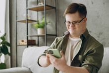 Young Man With Down Syndrome Wear Glasses Casual Clothes Use Mobile Cell Phone Chatting Sits On Grey Sofa Couch Stay At Home Flat Rest Spend Free Time In Living Room Genetic Disease World Day Concept