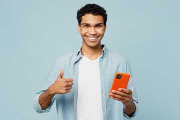 Wall Mural - Young smiling happy cheerful fun man of African American ethnicity he wears shirt casual clothes hold in hand use mobile cell phone show thumb up isolated on plain pastel light blue cyan background.