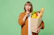 Elderly woman wear brown shirt casual clothes hold shopping paper bag with food products say hush be quiet finger on lips shhh gesture isolated on plain green background. Delivery service from shop.