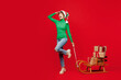 © ViDi Studio - Full body young woman wear green turtleneck Santa hat posing carry many present boxes on sled look far away isolated on plain red background. Happy New Year 2024 celebration Christmas holiday concept.