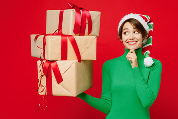 Wall Mural - Young fun minded woman wears green turtleneck Santa hat posing hold many present boxes with gift ribbon bow look aside isolated on plain red background Happy New Year 2024 Christmas holiday concept.