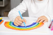 © Zarina Lukash - A small child draws a rainbow with felt-tip pens in a sketchbook while sitting at a table