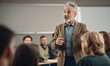 © Andrii IURLOV - Professor, Teacher Man 55 years old with grey hair giving a lecture at university  in big classroom at high school before many sitting students, Expert Does Motivational Talk.