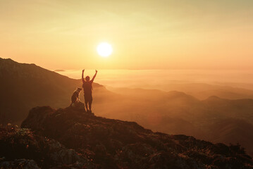 silhouette of a mountaineer man raising his arms and contemplating the sunset after hiking in the mountains with his border collie breed dog. outdoor sports and adventure