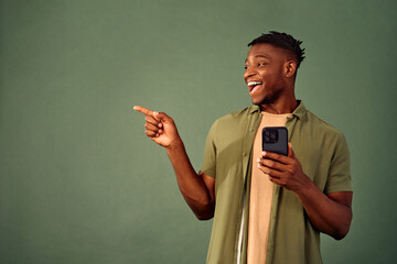 Wall Mural - Check this new app. Attractive african american young guy pointing aside with happy facial expression while holding modern smartphone in hand. Copy space for advertising on green studio background.