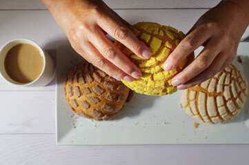 Wall Mural - Hands of Mexican Hispanic woman. Concept of taking food with hands or handling food. Taking a 
Mexican sweet bread concha with two hands, in the bottom conchas on a white plate and coffee