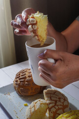 Wall Mural - Hands of Mexican Hispanic woman. Concept of taking food with hands or handling food. Soaking a piece of sweet bread in a cup of coffee over a plate with conchas near a kitchen window