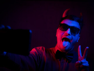 brunette man in a red shirt with a black bow tie in a black hat with red horns as a devil  make selfies  on a dark background, red and blue lighting is cinematic