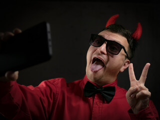 Wall Mural - brunette man in a red shirt with a black bow tie in a black hat with red horns as a devil on a dark background cinematic lighting