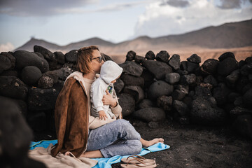 Wall Mural - Mother enjoying winter vacations playing with his infant baby boy son on black sandy volcanic beach of Janubio on Lanzarote island, Spain on windy overcast day. Family travel vacations concept