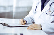 © rogerphoto - Unknown male doctor sitting and working with clipboard of medication history record in clinic at his working place, close-up. Young physician at work. Perfect medical service, medicine concept