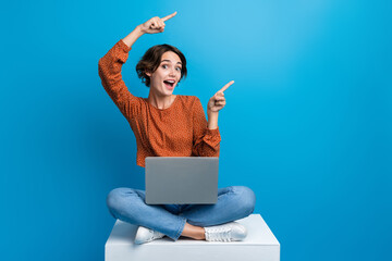 Full length photo of lovely young lady sit cube point empty space netbook wear trendy brown garment isolated on blue color background