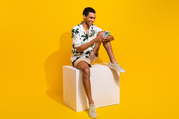 Wall Mural - Full size photo of handsome young guy sit white podium hold device wear trendy palms print outfit isolated on yellow color background