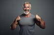 Middle aged grey haired man with standing with arms showing muscles happy smiling on camera wearing grey t-shirt isolated on grey background. Mature fit man, healthy lifestyle concept. Copy space 