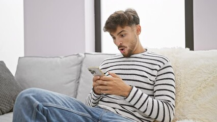 Wall Mural - Surprised young arab man pointing at smartphone while joyfully sitting on sofa at home, face glowing with amazement and positive expression.