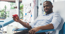 Black Businessman Donating Blood For People In Need. African Man Squeezing Heart-Shaped Red Ball To Pump Blood Through The Tubing Into Bag And Showing It To Camera As A Symbol Of Support And Smiling.
