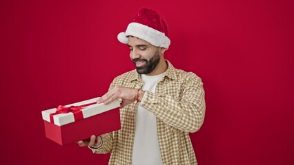 Sticker - Young hispanic man wearing christmas hat unpacking gift looking upset over isolated red background