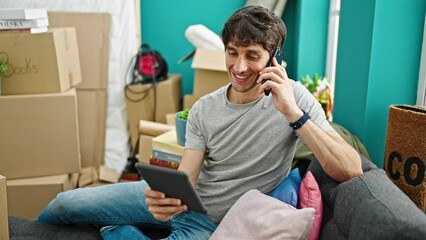 Wall Mural - Young hispanic man talking on smartphone using touchpad sitting on sofa at new home