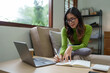 © Phimwilai - Young successful professional Asian businesswoman in casual clothes sits on a sofa, reads a book, writes notes in a notebook while working on a laptop computer at home office in the living room.