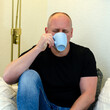 © dimas830 - An adult man drinks tea while sitting on a sofa in a room