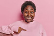 Positive dark skinned obese curly woman points index finger at herself asks who me and smiles gladfully being picked or chosen wears casual jumper isolated over pink background. Do you mean me
