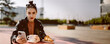 A beautiful young brunette woman sitting at a street café with coffee and a croissant, she is having a breakfast. She holds a smartphone in her hands. Panoramic banner picture with copy space.