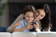Close Up Smiling Asian Mother With Caucasian Son Using Phone Together, Looking At Device Screen, Making Video Call Or Watching Cartoons, Hugging, Spending Leisure Time With Smartphone At Home