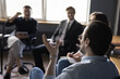 © fizkes - Group of office workers sit in circle at meeting in office discuss business idea, engaged in teambuilding activity. Confident male leader explain new strategies to creative team. Teamwork, cooperation