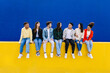 Young group of diverse friends having fun while hanging out sitting on yellow wall over blue background. Copy space for text. Youth community and friendship concept.