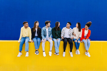 Wall Mural - Young group of diverse friends having fun while hanging out sitting on yellow wall over blue background. Copy space for text. Youth community and friendship concept.