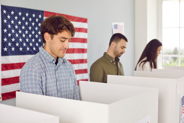 Wall Mural - American citizens voting at a polling station during presidential elections in the USA. Group of young men and women in voter booths at the ballot station with the US flag on the wall