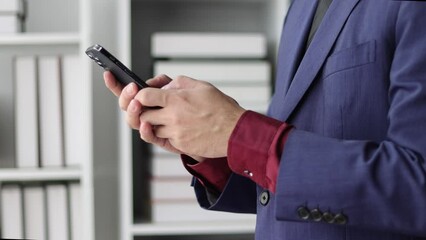 A businessman holding a smartphone is using the mobile phone to send messages online and is typing.