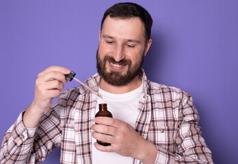 Wall Mural - Handsome bearded man holding pipette with beard oil on purple background. 