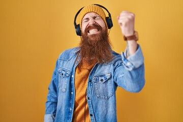 Wall Mural - Caucasian man with long beard listening to music using headphones angry and mad raising fist frustrated and furious while shouting with anger. rage and aggressive concept.