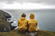 Young couple wearing yellow raincoats sitting on the edge of a cliff with huge waves rolling ashore. Rough Irish weather. Beautiful nature of Ireland.