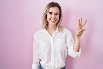 Poster - Young beautiful woman standing over pink background showing and pointing up with fingers number three while smiling confident and happy.