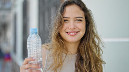 Wall Mural - Young beautiful hispanic woman holding bottle of water smiling at street
