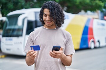 Wall Mural - Young latin man using smartphone and credit card at bus station