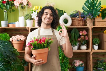 Sticker - Hispanic man with curly hair working at florist shop holding plant doing ok sign with fingers, smiling friendly gesturing excellent symbol