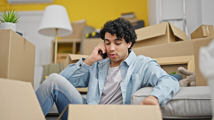 Sticker - Young latin man talking on smartphone unpacking cardboard box looking upset at new home