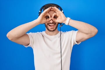 Poster - Hispanic man with beard listening to music wearing headphones doing ok gesture like binoculars sticking tongue out, eyes looking through fingers. crazy expression.