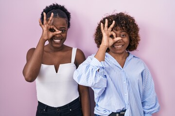 Wall Mural - Two african women standing over pink background doing ok gesture with hand smiling, eye looking through fingers with happy face.