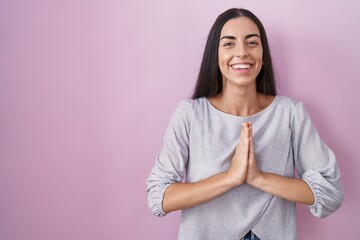 Poster - Young brunette woman standing over pink background praying with hands together asking for forgiveness smiling confident.