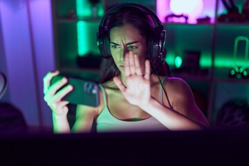 Poster - Young brunette woman playing video games with smartphone with open hand doing stop sign with serious and confident expression, defense gesture