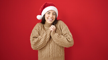 Wall Mural - Young beautiful hispanic woman smiling confident wearing christmas hat over isolated red background