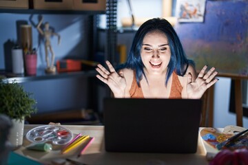 Wall Mural - Young modern girl with blue hair sitting at art studio with laptop at night celebrating crazy and amazed for success with arms raised and open eyes screaming excited. winner concept