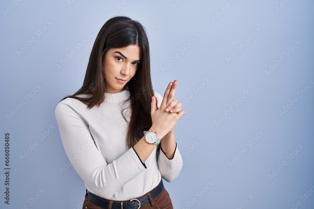 Young brunette woman standing over blue background holding symbolic gun with hand gesture, playing killing shooting weapons, angry face