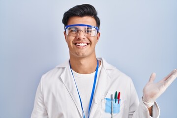 Poster - Hispanic man working as scientist smiling cheerful presenting and pointing with palm of hand looking at the camera.