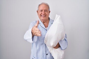 Poster - Senior man with grey hair wearing pijama hugging pillow smiling friendly offering handshake as greeting and welcoming. successful business.