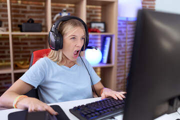 Poster - Young caucasian woman playing video games wearing headphones in shock face, looking skeptical and sarcastic, surprised with open mouth